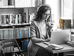 A woman is sitting at a desk using a laptop computer and drinking coffee.