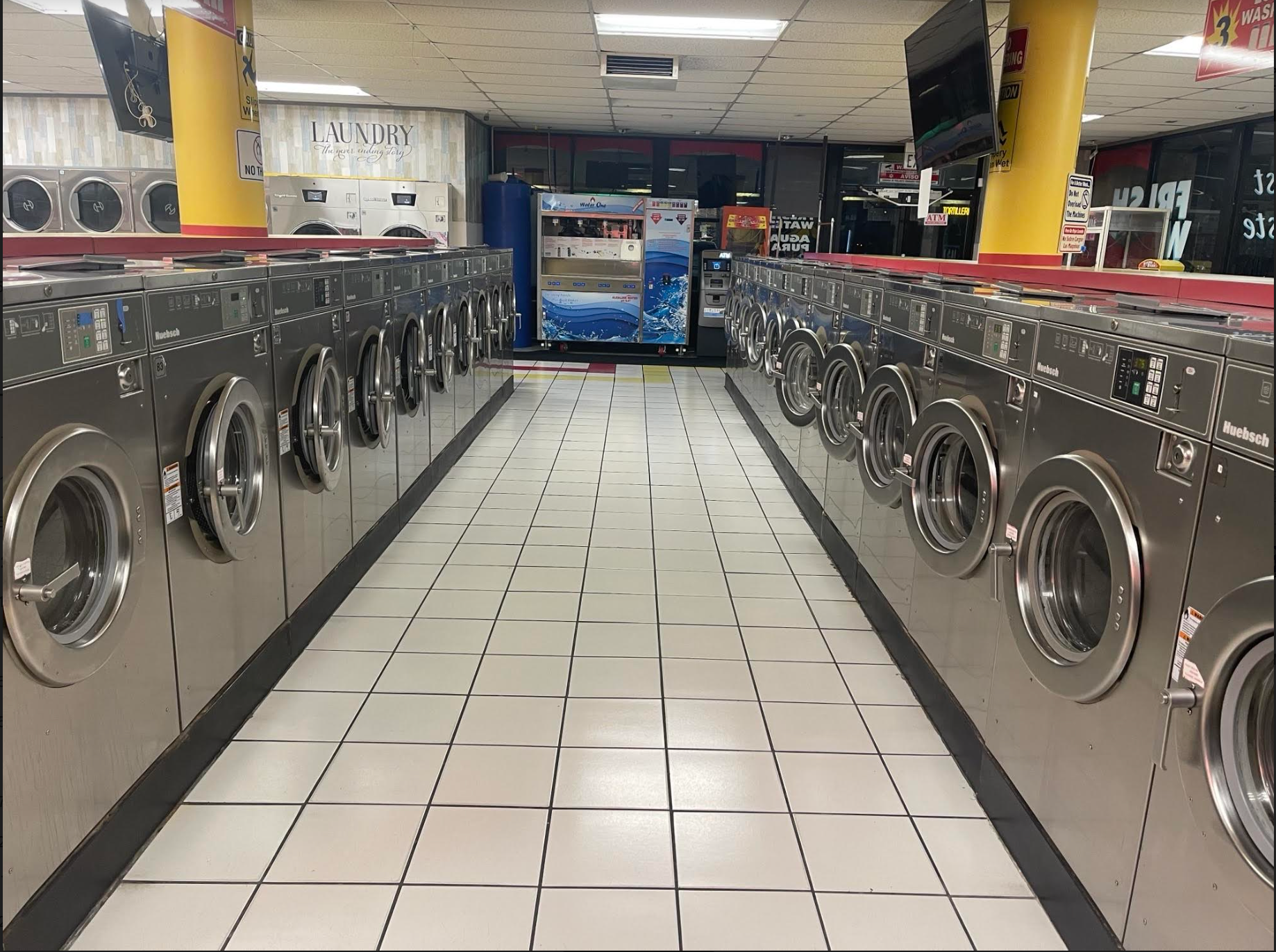 Rows of washing machines in a laundromat, with tile floor and a vending machine visible in the background.