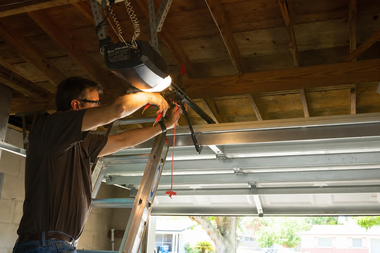 Person repairing a garage door opener under a wooden ceiling with a ladder and bright work light
