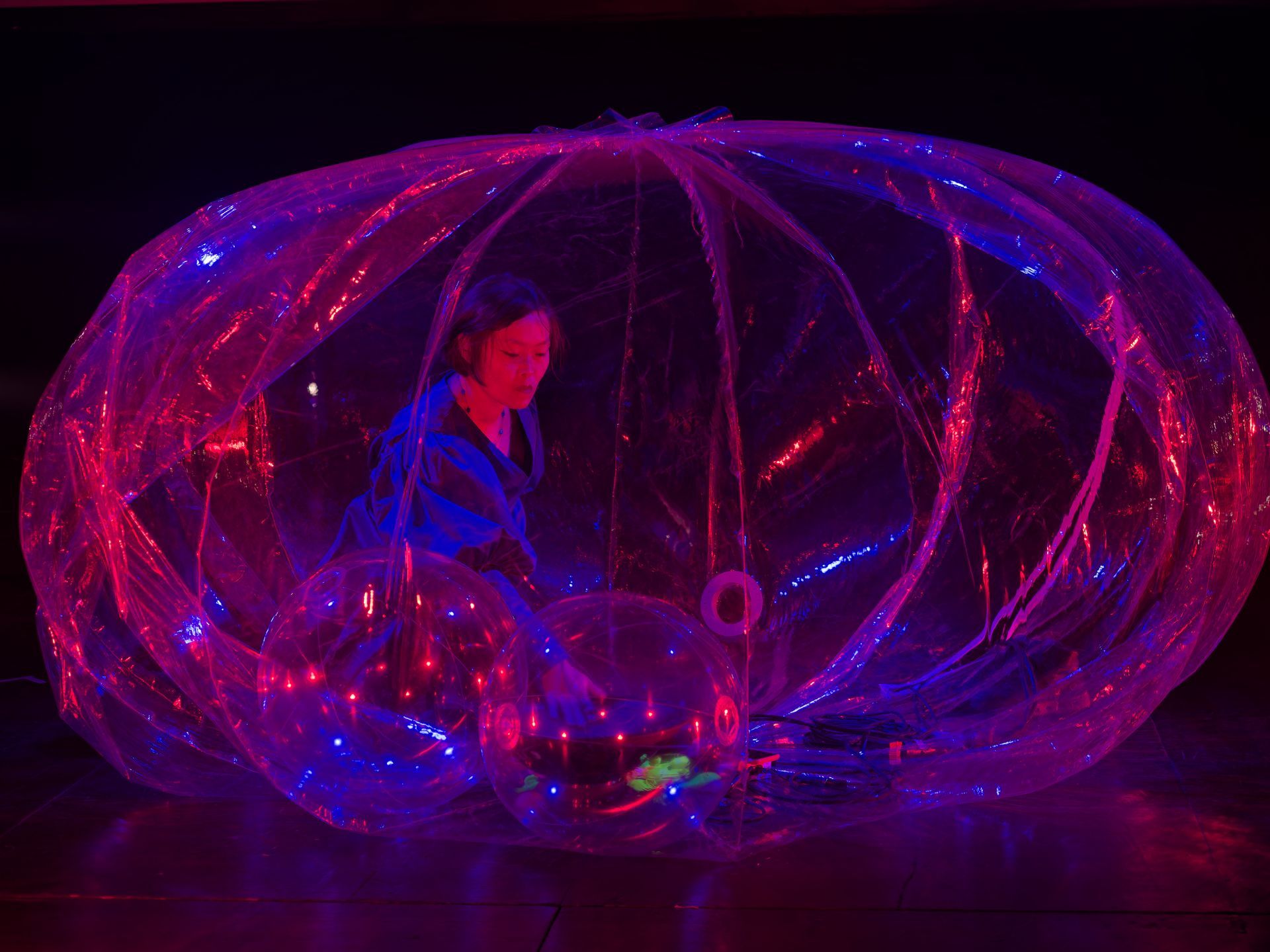 A performer in a blue jacket sits inside a large, transparent bubble, reaching upward amidst glowing purple lights.