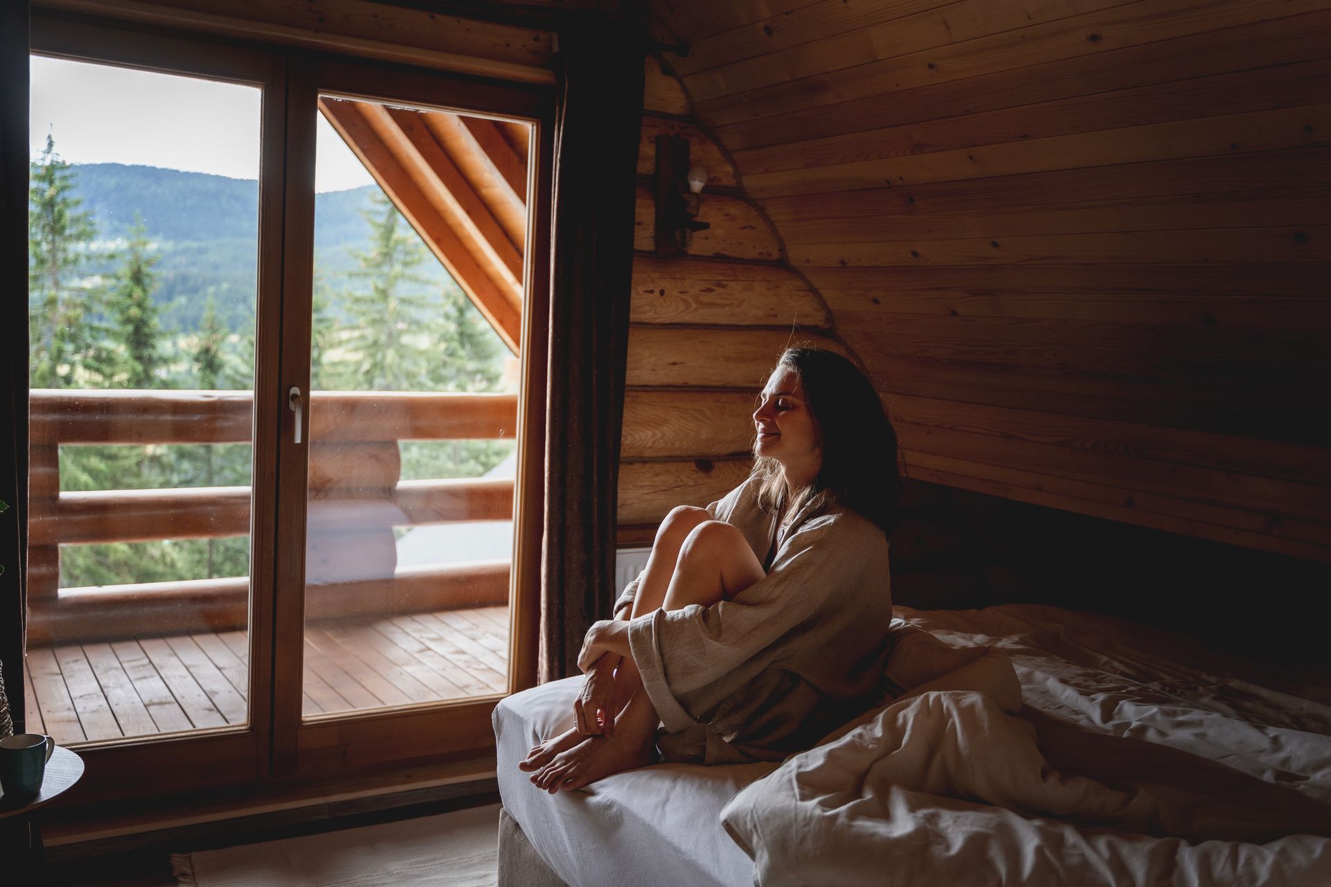 A person sits on a bed in a rustic wooden cabin, looking out a large glass door at a mountain view with pine trees.