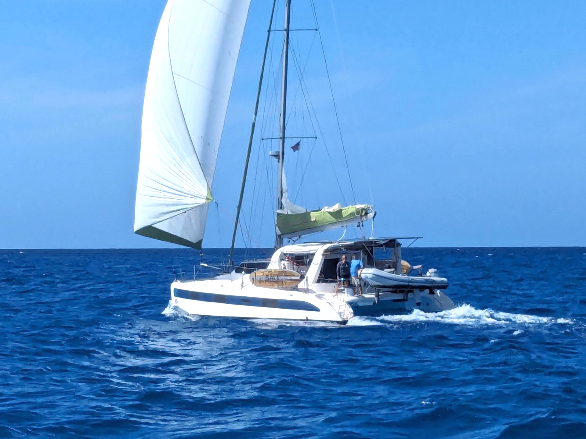A sailboat with a white sail travels across deep blue, calm ocean water, viewed from directly above.