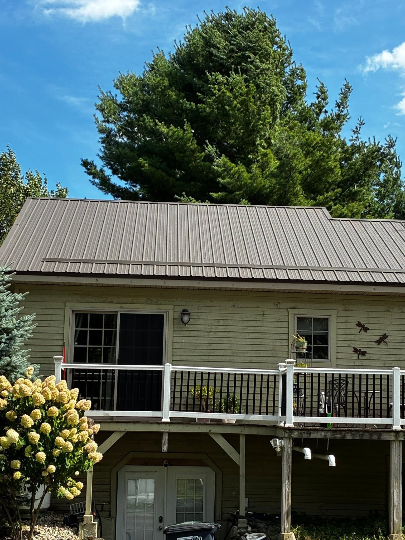 Two-story house with beige siding, gray roof, and white balcony railing, surrounded by trees under a blue sky