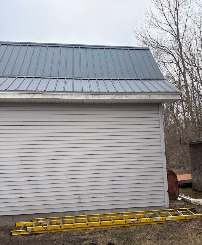 Gray-sided building with a metal roof and a yellow ladder lying on the ground beside it