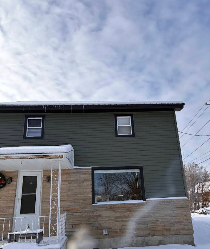 Snow-covered two-story house with green siding, tan stone base, and cloudy winter sky