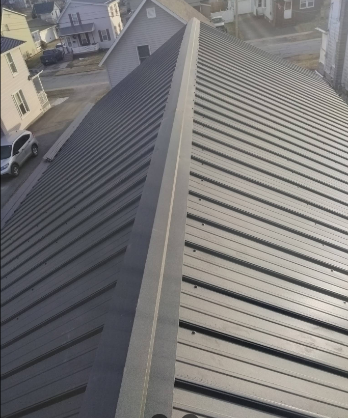 Rooftop view of a dark metal roof with ridges and neighboring houses in the background