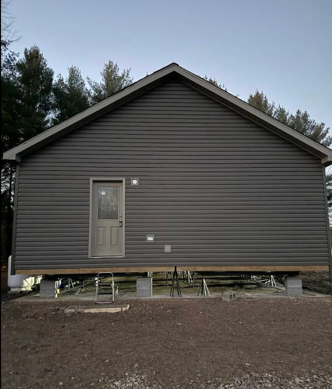 Small gray house exterior with a gable roof and front door, set on raised supports over bare ground.