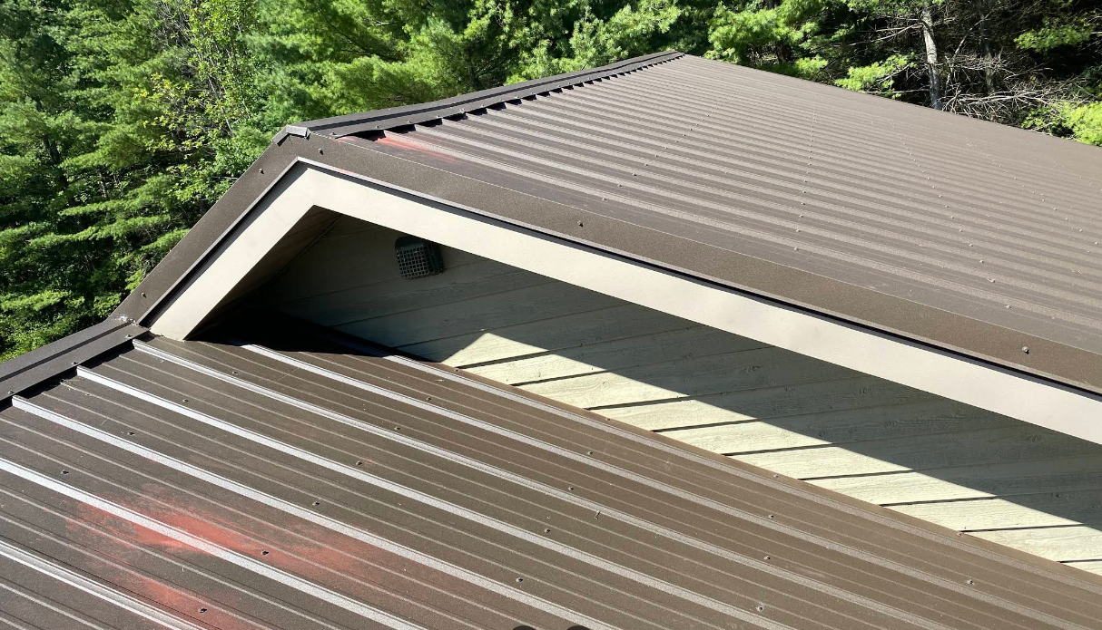 Metal-roofed building roof with a triangular vent, surrounded by green trees