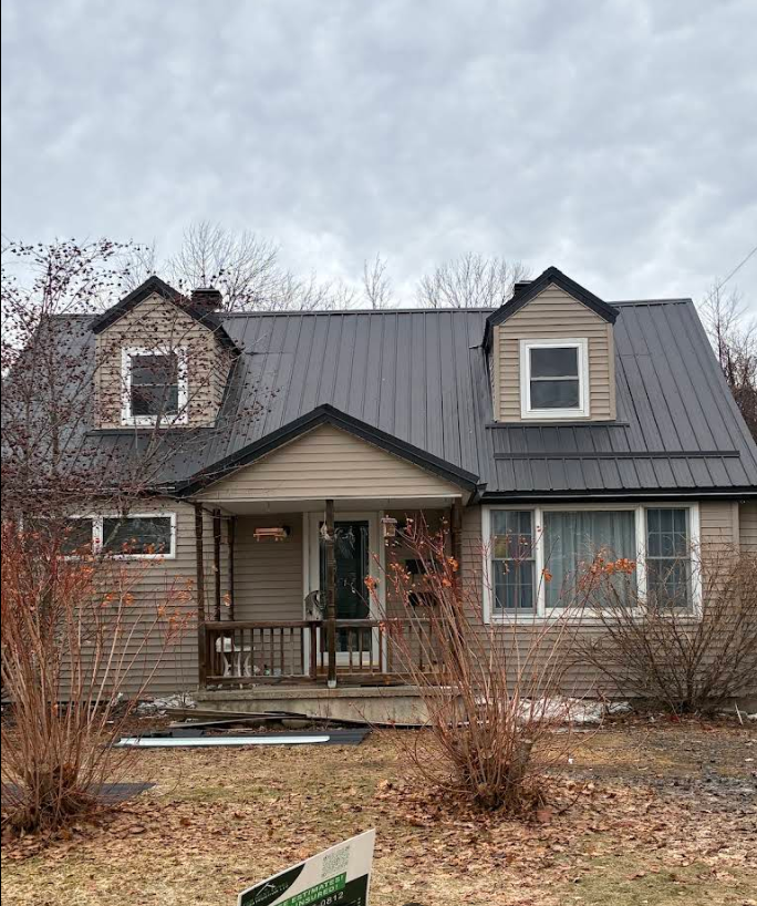 Small beige house with dark gray roof, front porch, bare shrubs, and cloudy sky.