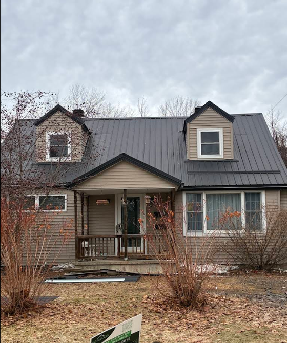 Two-story beige house with a wraparound deck and tan roof, surrounded by trees and shrubs.