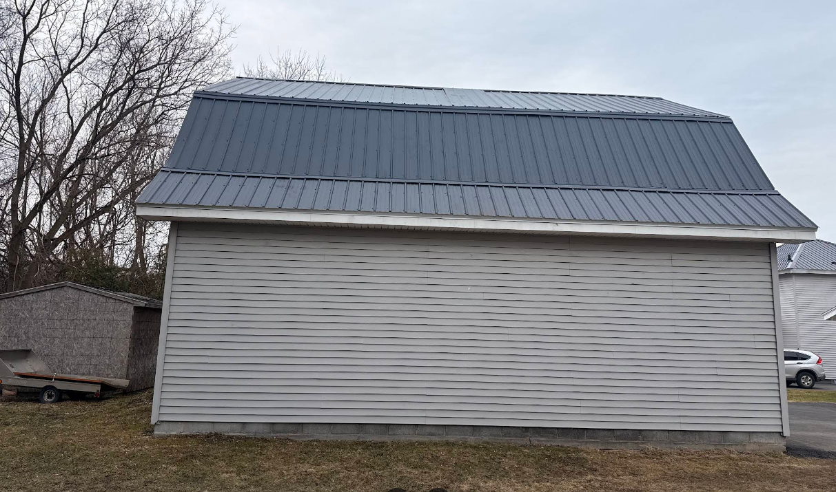 Small white shed with a gray metal roof in a rural yard, with bare trees and overcast sky.