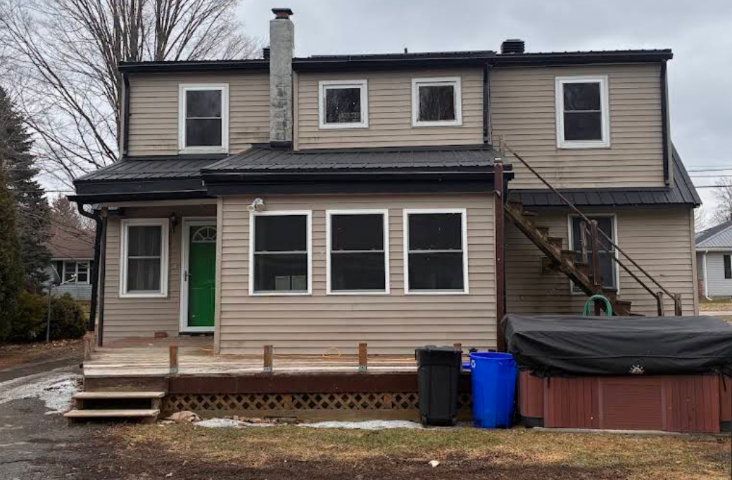 Two-story beige house with green door, front porch, stairs, and blue trash bins