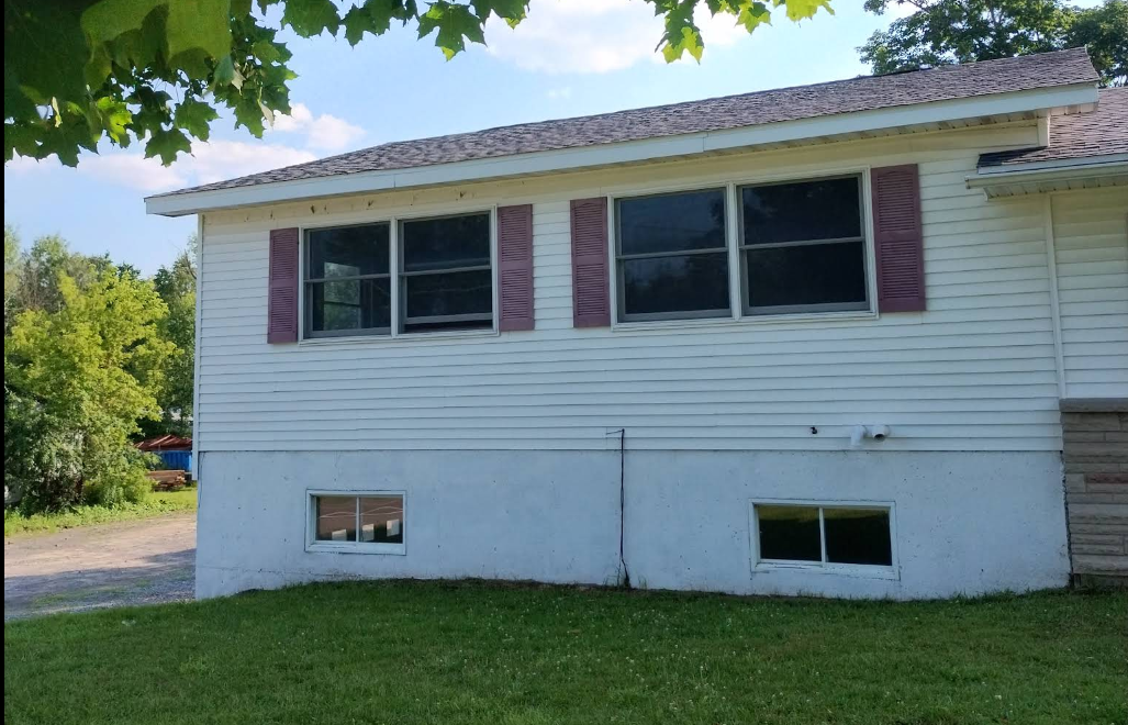 Side view of a white house with burgundy shutters, small basement windows, and a green lawn under trees.