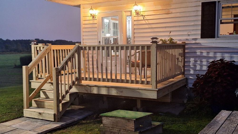 Small wooden porch with stairs and railing lit by a porch light at dusk beside a house.