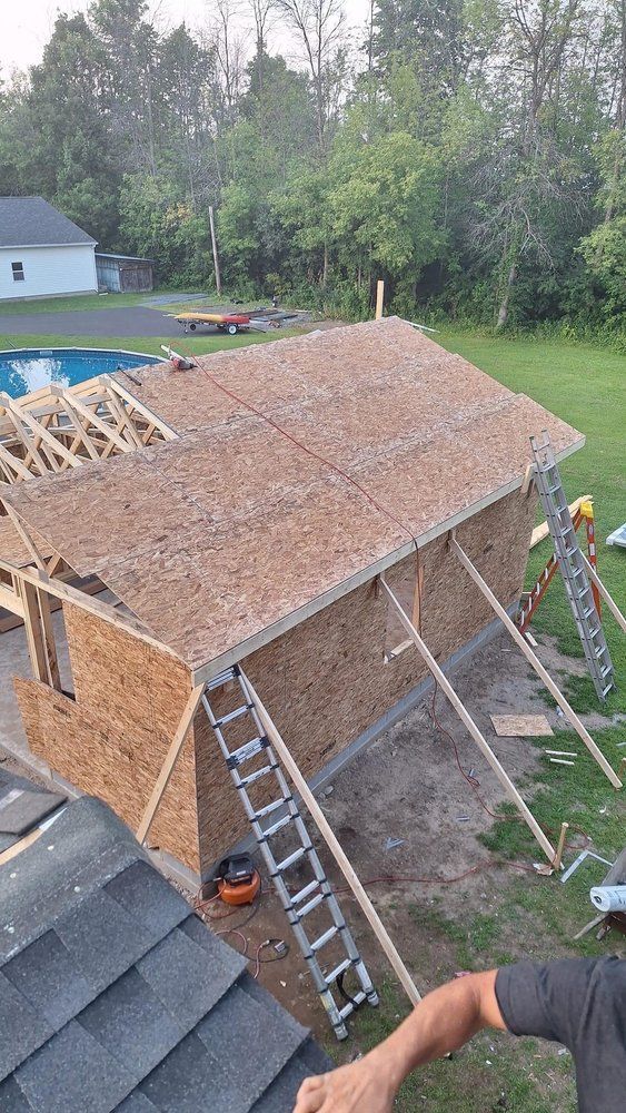 Workers framing a small wooden roof on a shed, with ladders and tools at a construction site.
