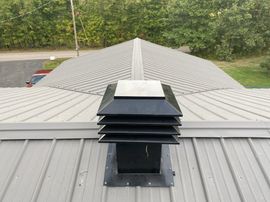 Metal roof vent centered on a gray shingled rooftop with trees in the background