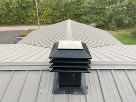 Metal roof vent centered on a gray shingled rooftop with trees in the background