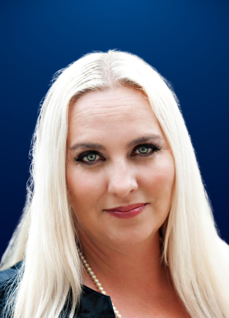 A woman with long blonde hair is wearing a black jacket and pearls.
