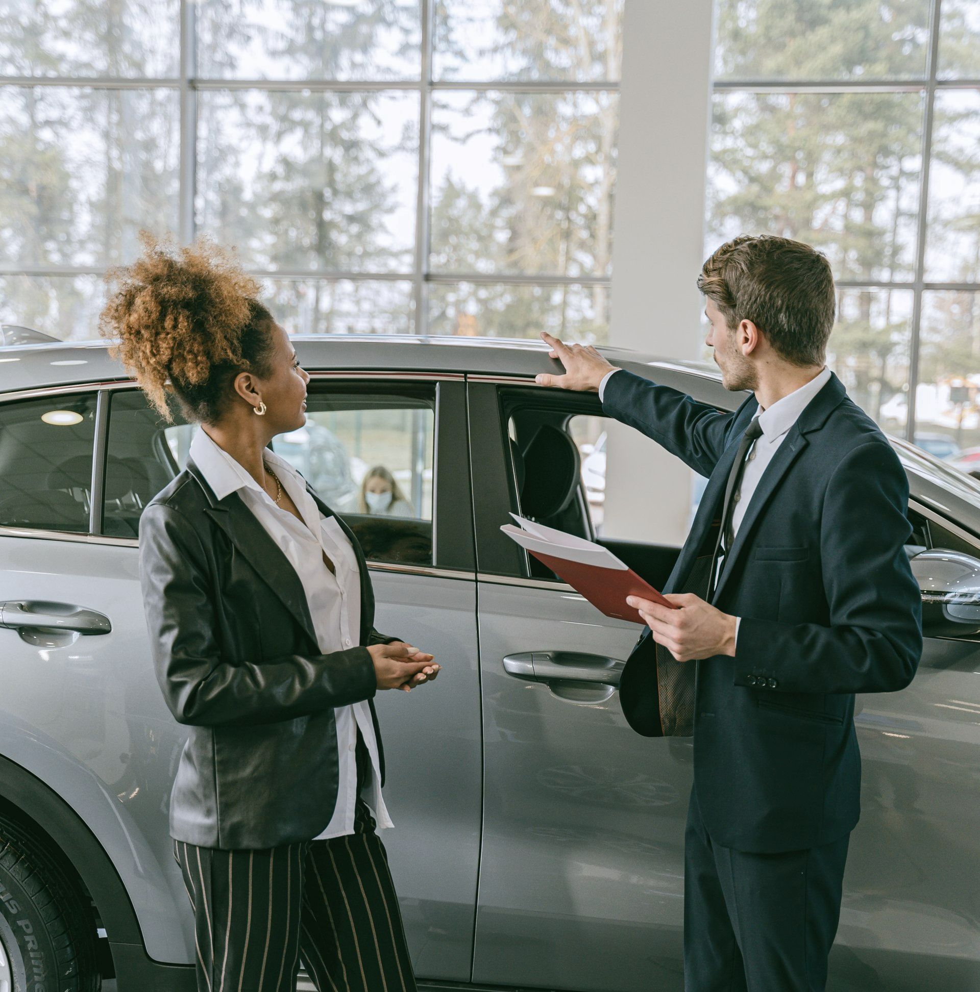Two businesspeople discussing a silver car in a bright showroom, one holding a clipboard.
