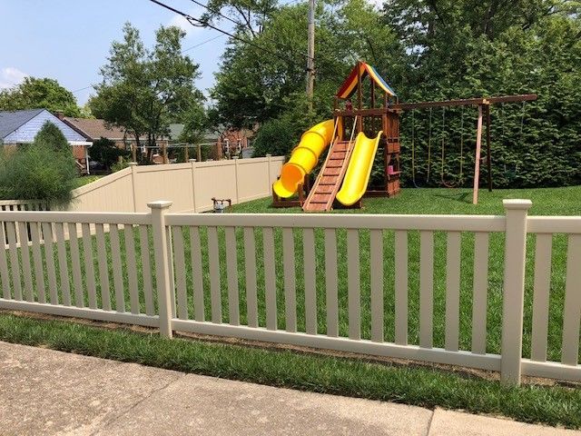 A white fence surrounds a playground with a yellow slide.