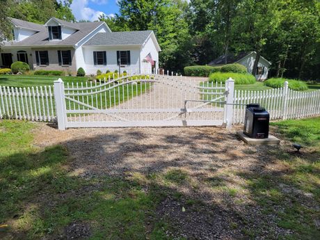 White picket gate and fence in front of a white house with a gravel driveway and lawn.