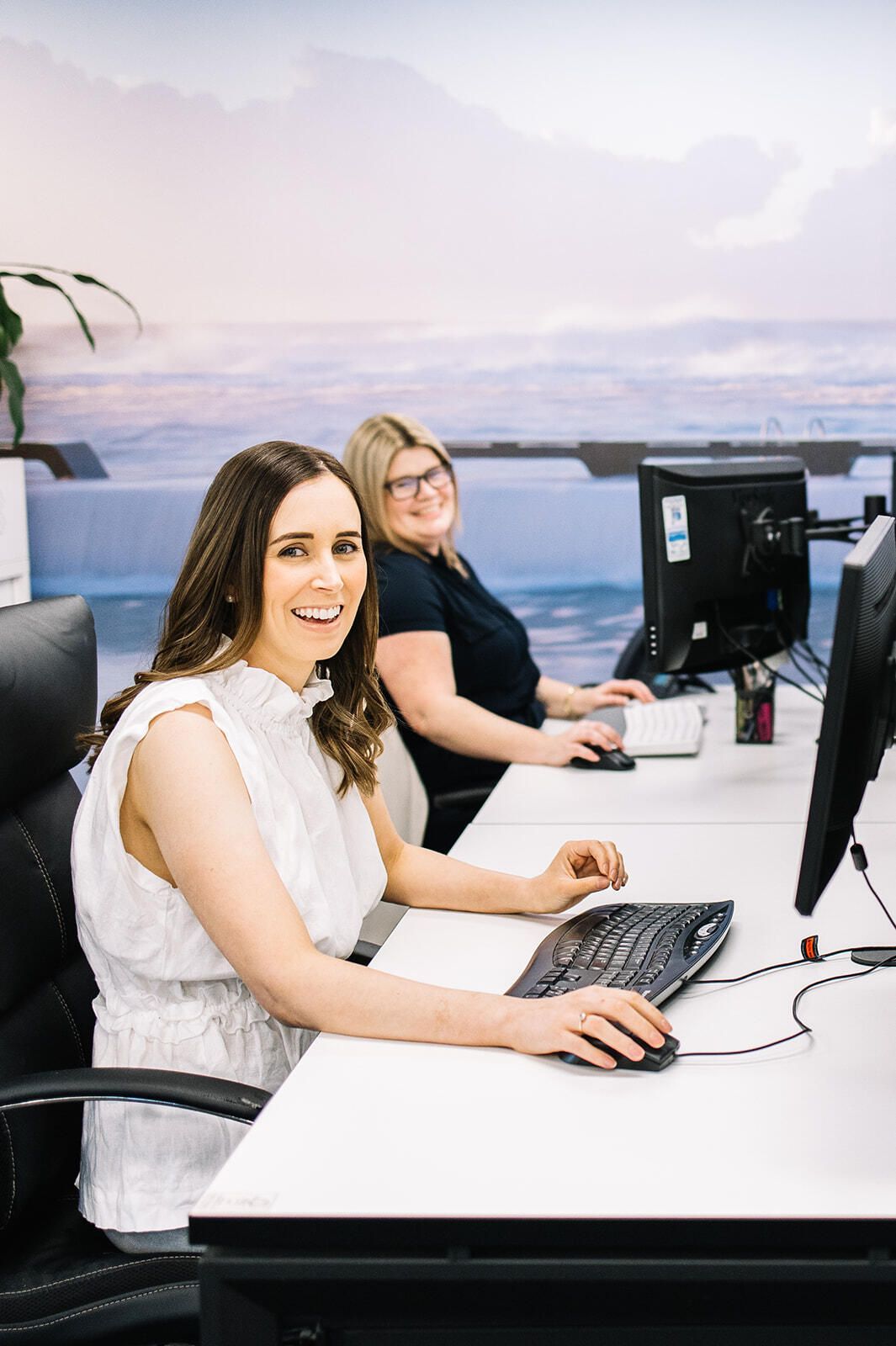 Two Women Are Sitting At Desks In Front Of Computer Monitors — The Garis Group  In Sydney, NSW