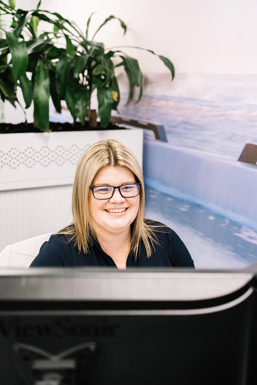 A Woman Wearing Glasses Is Smiling While Sitting In Front Of A Computer  Monitor — The Garis Group  In Sydney, NSW