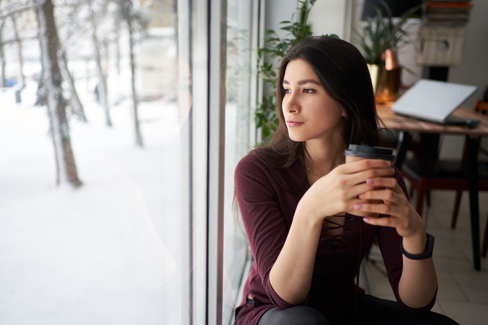 Woman by a snowy window holding a coffee mug, looking outside in a cozy café.