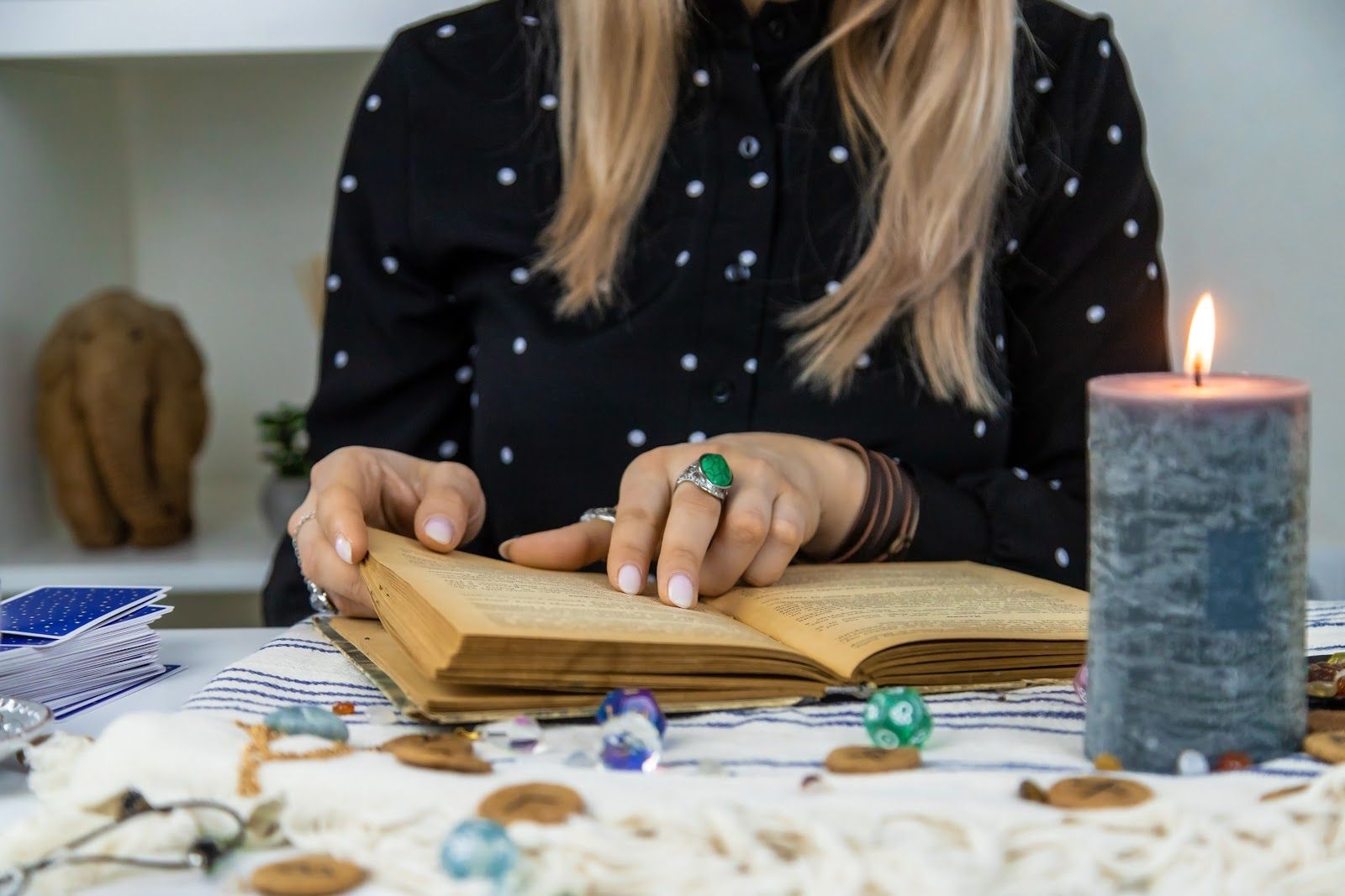 Hands open a book beside a lit candle, crystals, and tarot cards on a table