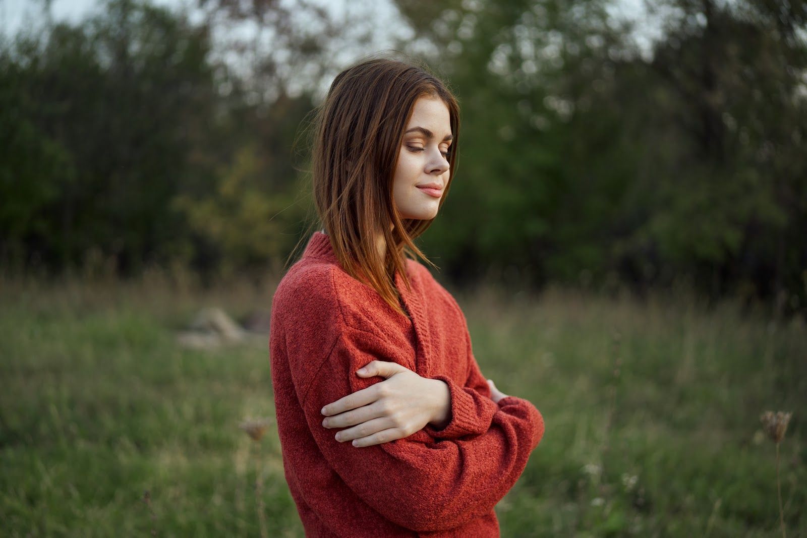 Person in a red sweater standing in a grassy field, arms crossed, with a calm expression
