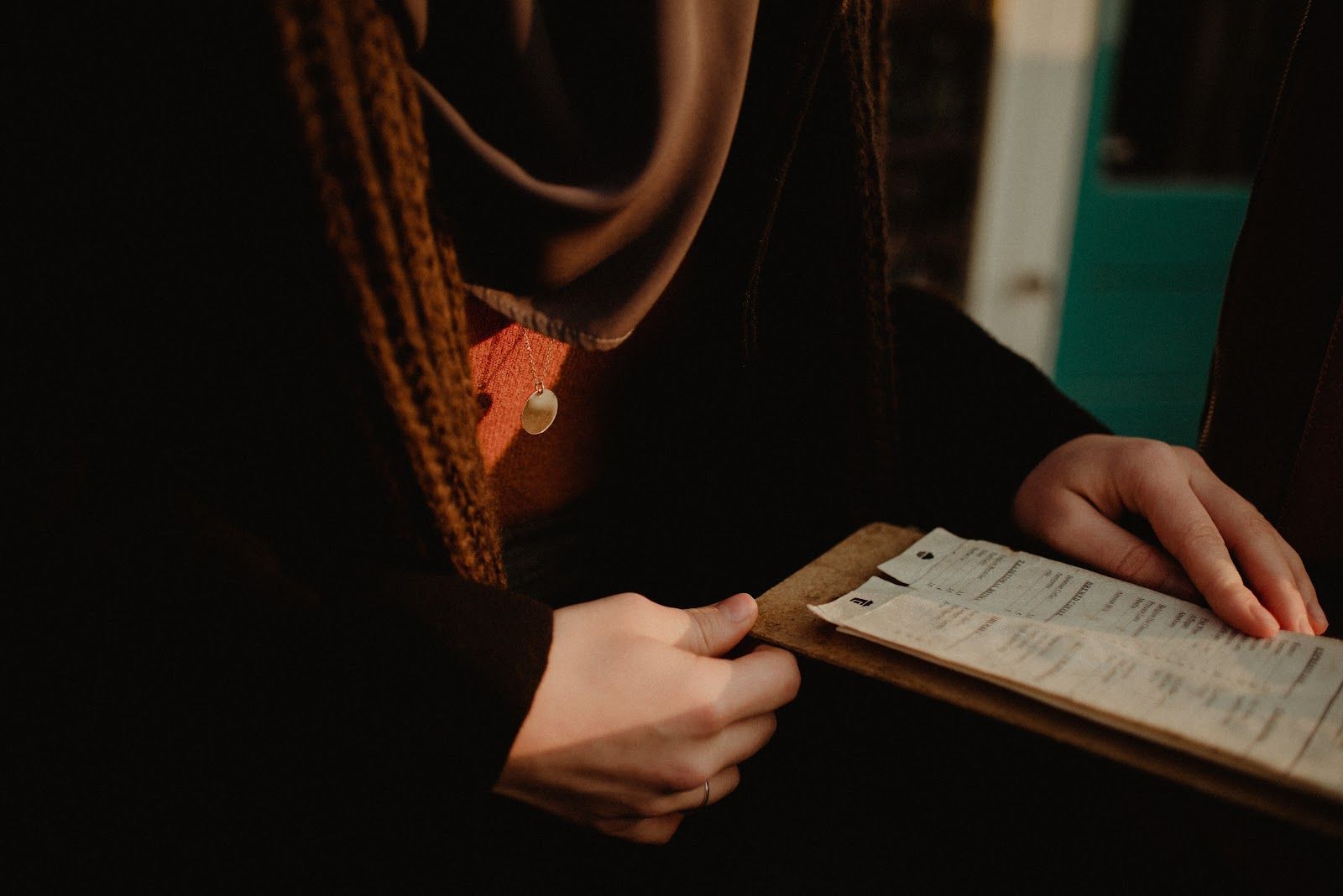 Hands holding an open notebook beside a table, with a dark scarf and sweater in warm light.