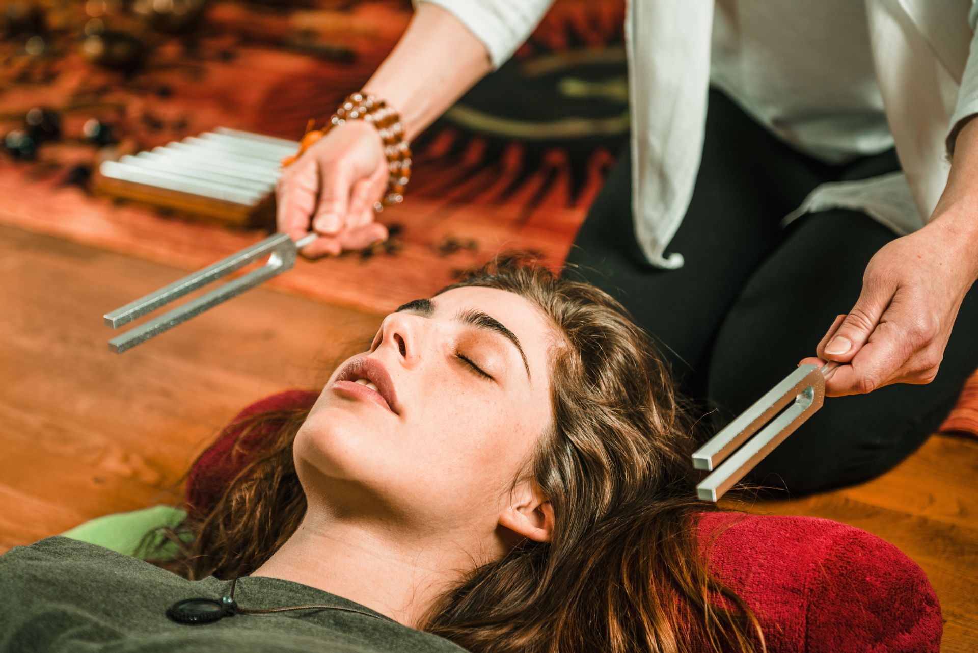A woman is laying on the floor while a person holds a pair of tuning forks over her head.