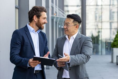 Two men in suits, conversing and gesturing, near a modern building's entrance. One holds a clipboard.