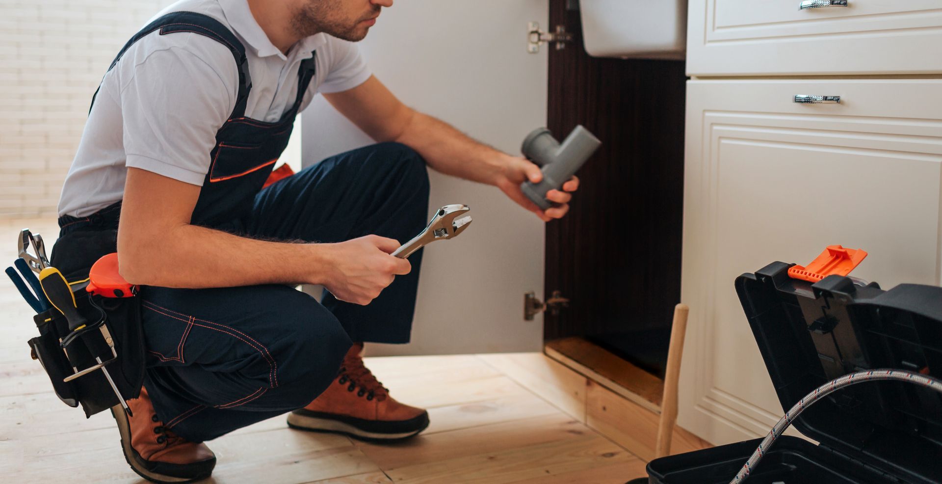Plumber kneeling in kitchen cabinet area, using tools to repair plumbing under the sink.
