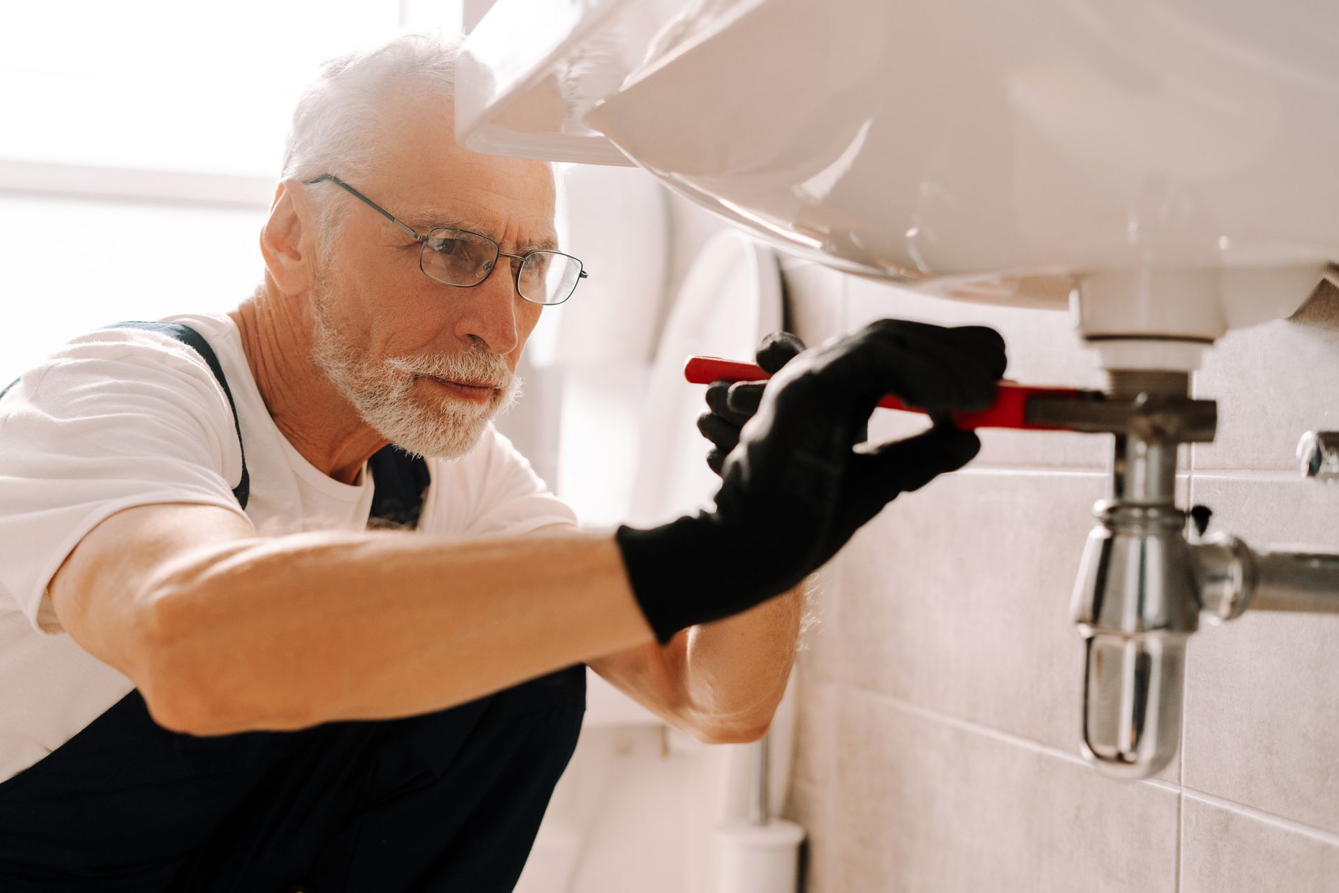 The plumber wearing gloves carefully fixes the sink pipe with adjustable pliers. Plumber kneels under a sink, using a wrench, wearing glasses and gloves.