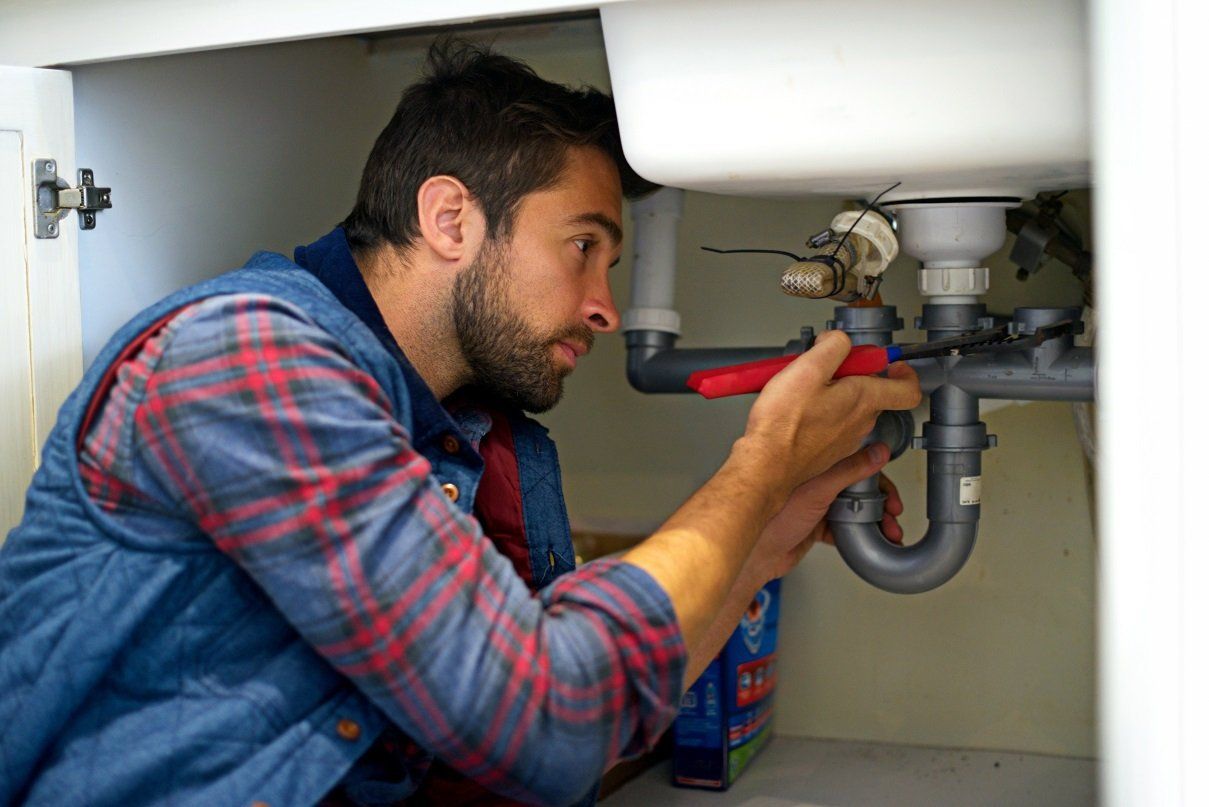 A plumber with a beard repairs pipes under a white sink in a cabinet.