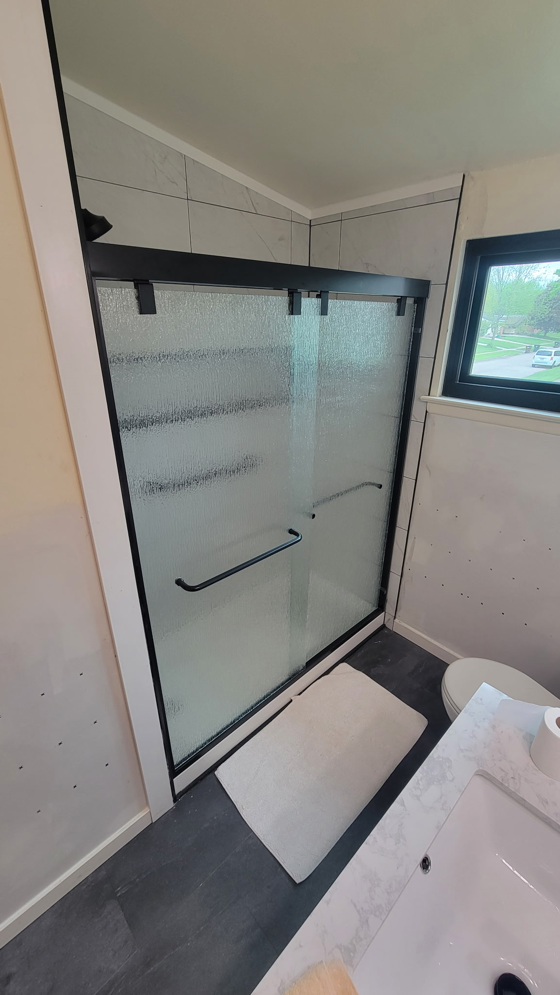 Bathroom with a shower, toilet, and sink. Black framed shower with frosted glass, white bath mat, and small window.