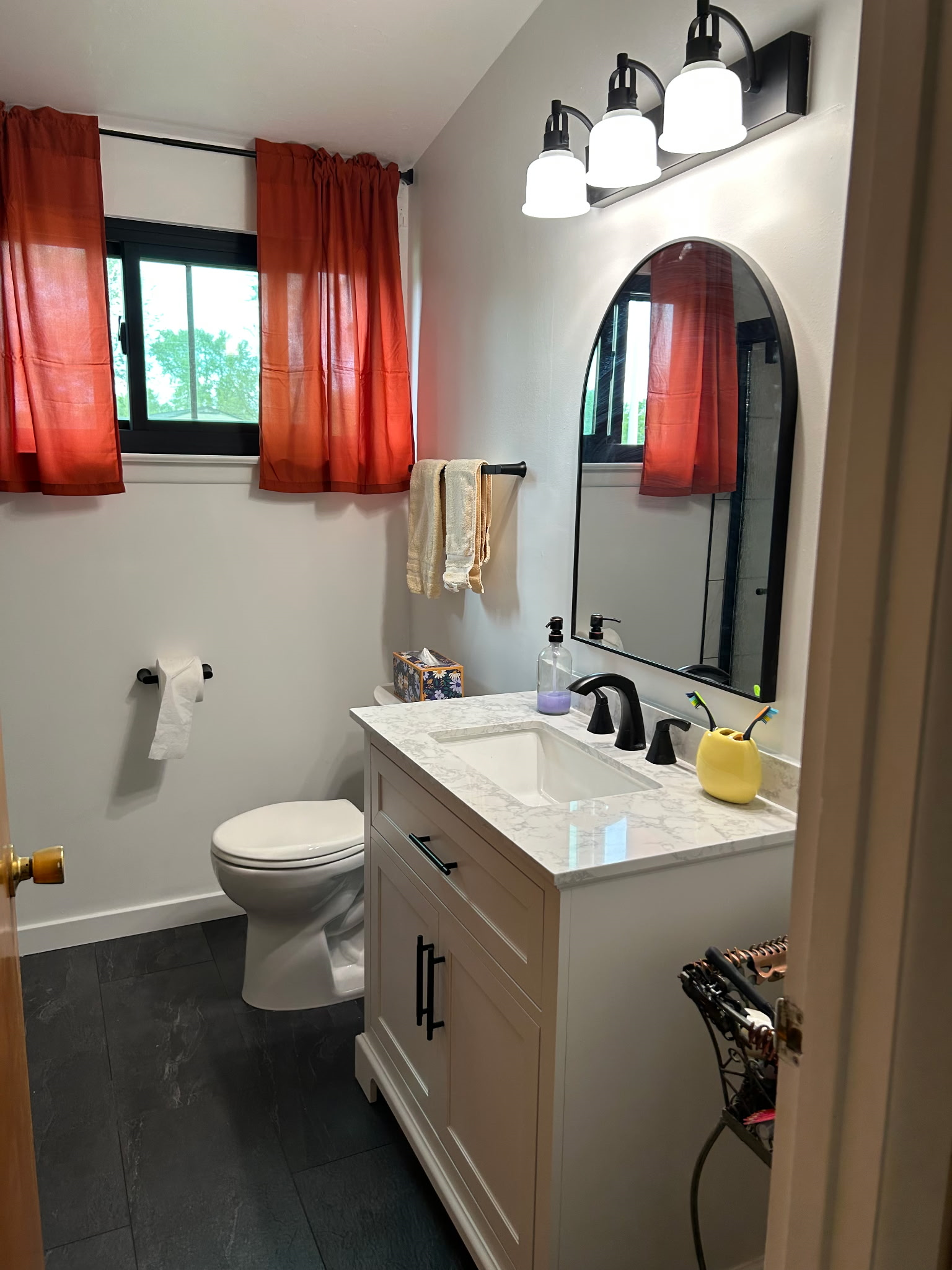 Bathroom with white vanity, black fixtures, arched mirror, orange curtains, and dark gray floor.
