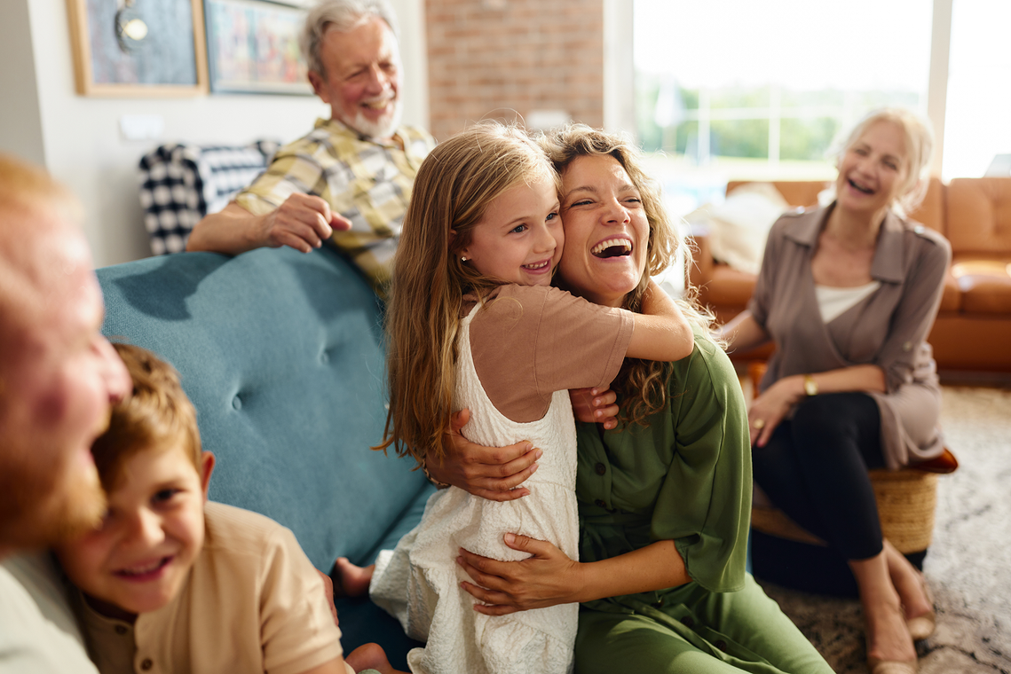 A family sits in a living room, smiling and laughing as a child embraces a woman on a blue sofa.
