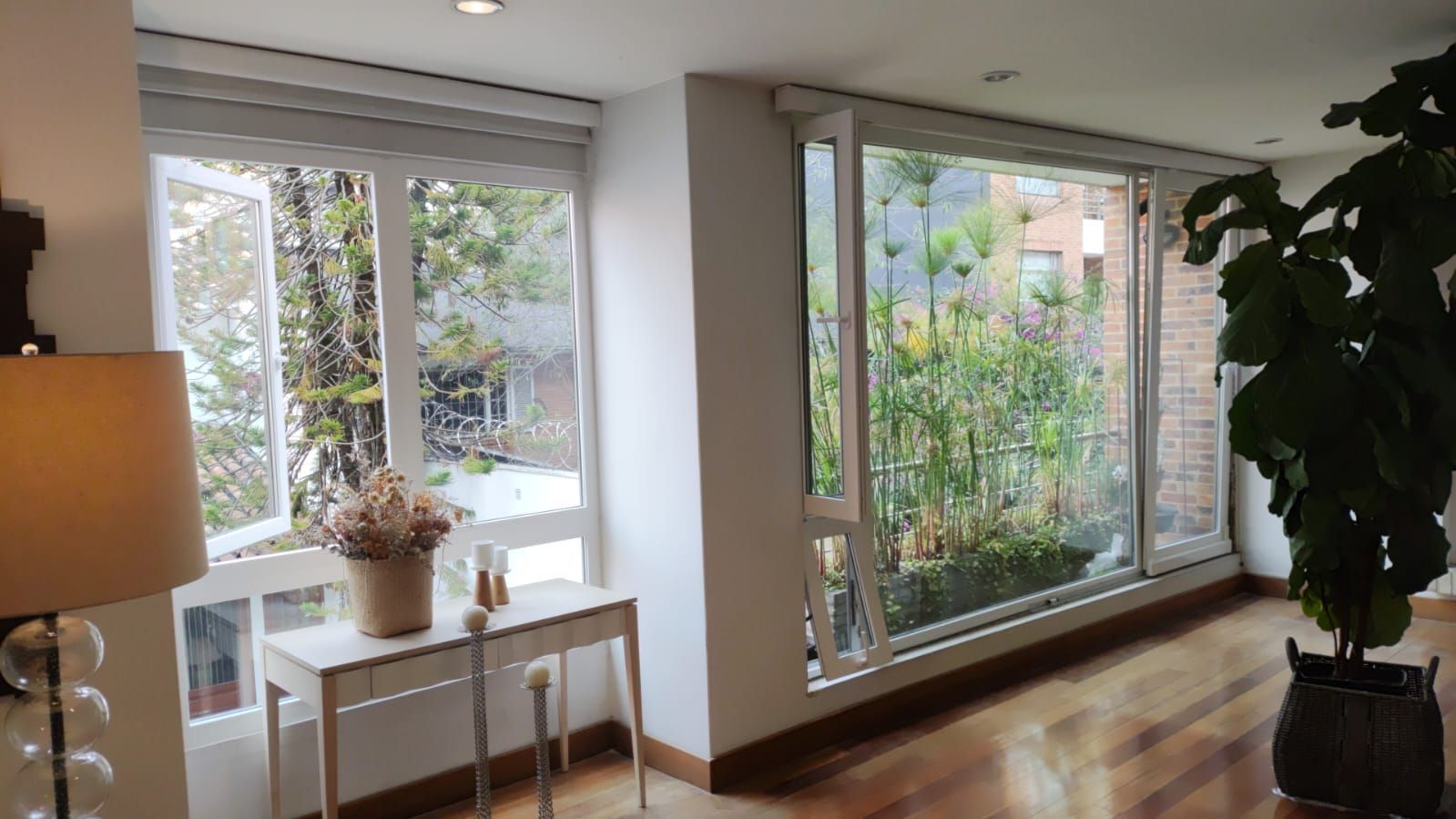 Sala de estar luminosa con grandes ventanales, paredes blancas, suelo de madera, lámpara, mesa y una planta alta en maceta.
