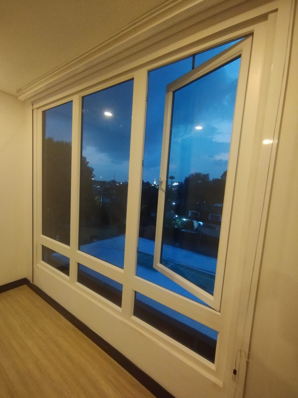Vista interior de una gran ventana con marco blanco al atardecer, que se abre a un paisaje urbano azul vespertino.