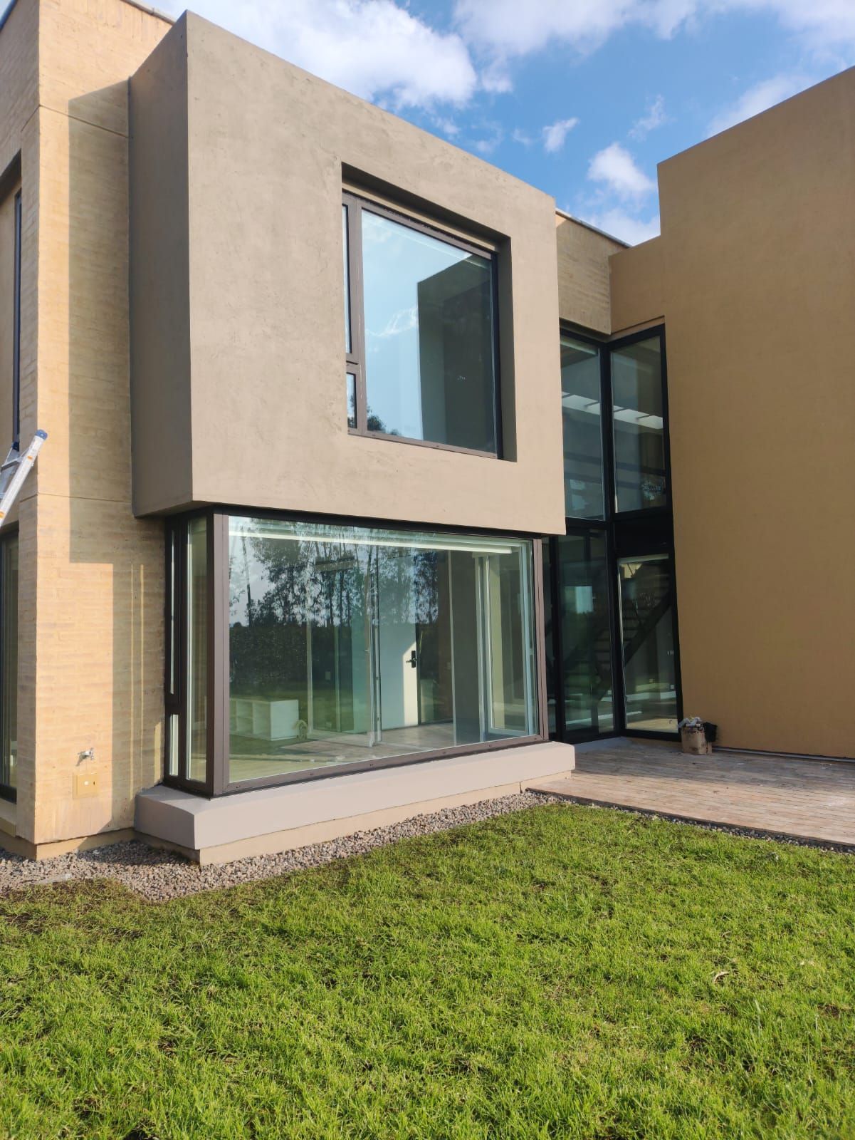 Exterior de casa moderna de color beige con grandes ventanales, entrada de esquina acristalada y césped verde.