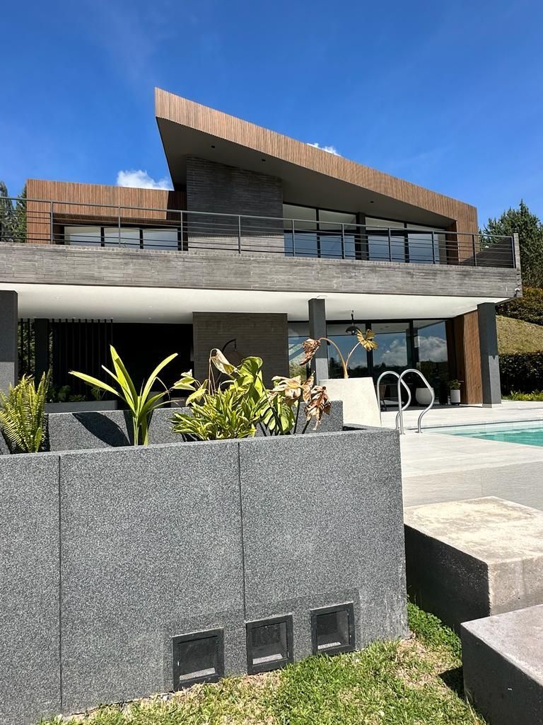 Exterior de una casa moderna con detalles en madera, balcón de cristal y jardinera de piedra gris bajo un cielo azul claro.
