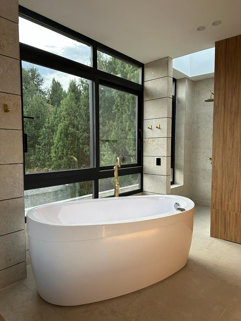Baño moderno con bañera ovalada blanca, ventanas con marcos negros y vistas al bosque natural.