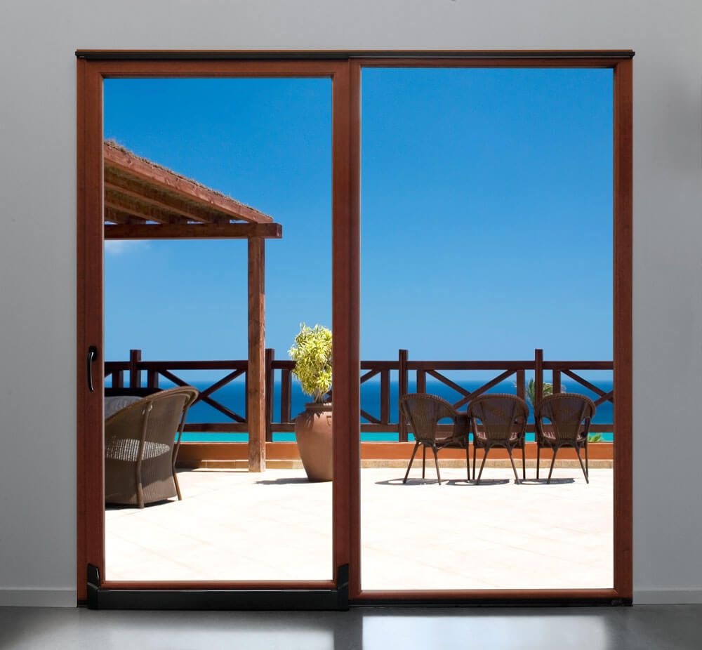 Balcón con vistas al mar a través de puertas corredizas de cristal, con sillas, mesa y cielo azul.