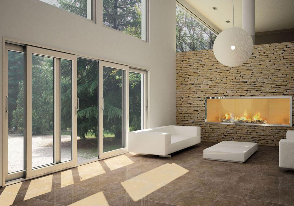 Salón moderno con grandes ventanales, sofás blancos, chimenea de piedra y luz solar cálida en el suelo.