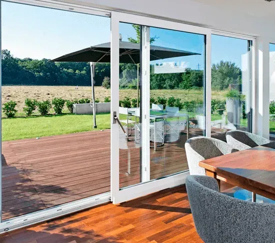 Unas puertas correderas de cristal dan acceso a una terraza de madera, un césped verde y una mesa de patio