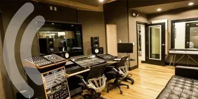 Sala de control de estudio de grabación con mesas de mezclas, sillas y cabinas de cristal insonorizadas.