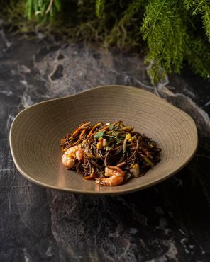 Noodles marroni con gamberi e verdure in una ciotola color sabbia dalla forma organica.