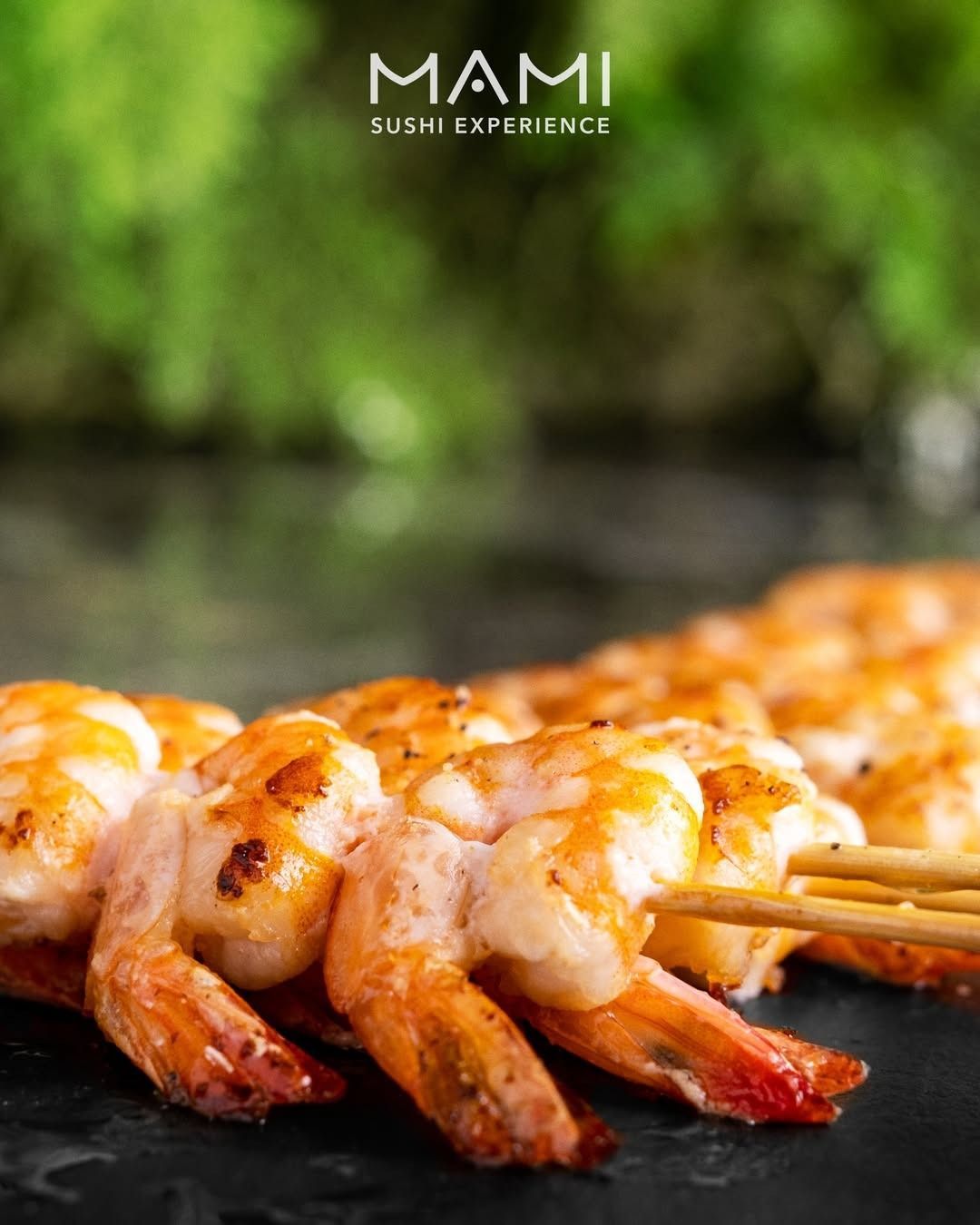 Gamberi grigliati su una superficie scura, con bacchette; logo MAMI Sushi Experience nell'angolo in alto.